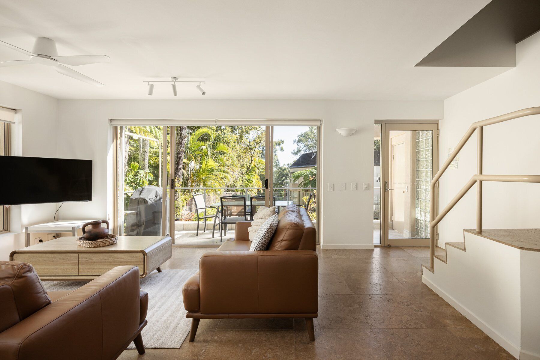 A modern living room with a flat-screen TV, leather sofas, a dining area with a table and chairs, and a sliding glass door leading to a balcony with a view of trees.