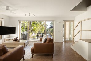 Una sala de estar moderna con un televisor de pantalla plana, sofás de cuero, un comedor con mesa y sillas, y un ventanal corredero que da a un balcón con vistas a árboles.