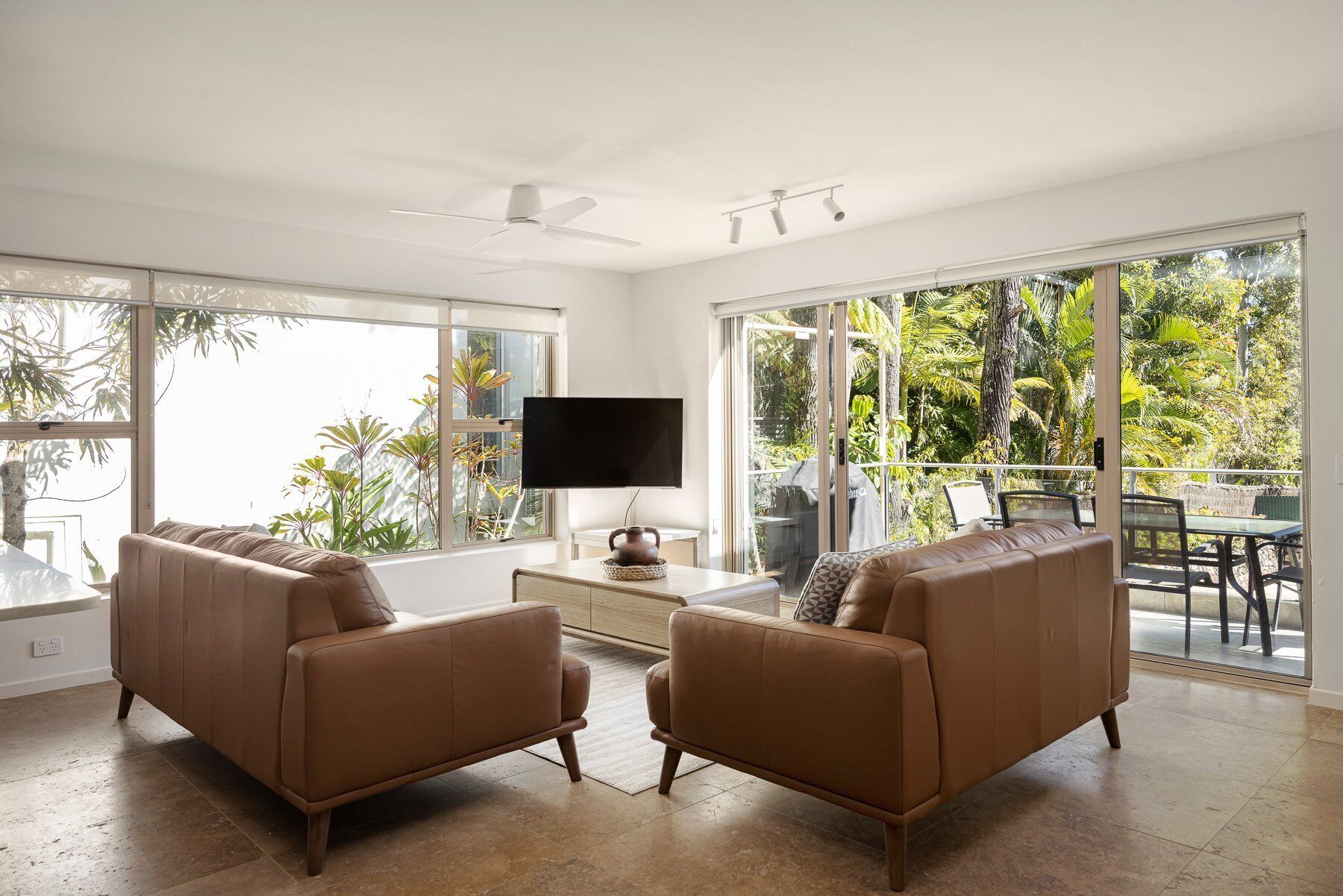 A spacious living room with two brown leather sofas, a flat-screen TV, and a ceiling fan. Large windows offer a view of lush greenery outside.