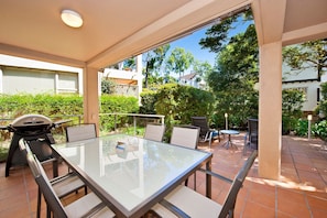 An outdoor patio with a glass table, chairs, a barbecue grill, and a view of a residential area.