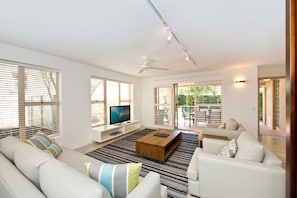 A modern living room with a flat-screen TV, white sofas, a coffee table, and a ceiling fan.