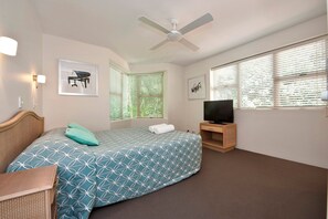A bedroom with a bed, a television, a ceiling fan, and two windows with blinds.