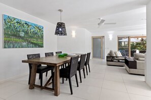 Un espace de salle à manger moderne avec une table et des chaises en bois, un luminaire suspendu et un tableau représentant une scène tropicale.