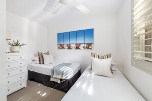 A bedroom with two beds, a dresser, a ceiling fan, and a beach-themed artwork on the wall.