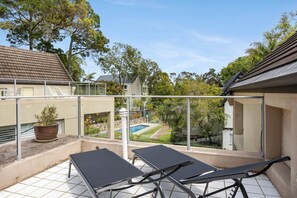 A balcony with two black lounge chairs, a potted plant, and a view of a swimming pool and garden.