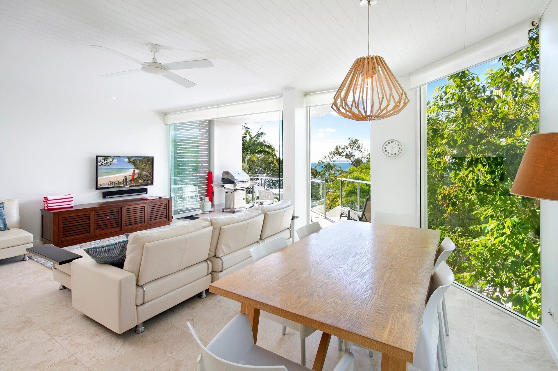 A modern living room with a large dining table, a sofa, a TV, and a balcony with a view of the ocean.