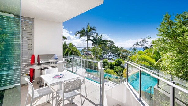 Un balcon avec une balustrade en verre, un barbecue, une table avec des chaises et une piscine.