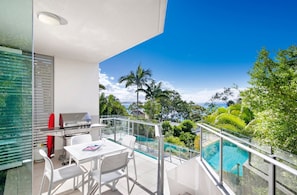 A balcony with a glass railing, a barbecue grill, a table with chairs, and a pool.