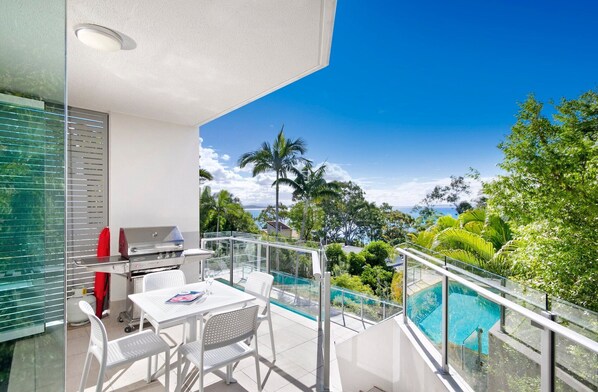 Un balcon avec une balustrade en verre, un barbecue, une table avec des chaises et une piscine.