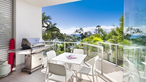 Un balcon équipé d’un barbecue, d’une table à manger, de chaises et offrant une vue sur l’océan et la végétation.