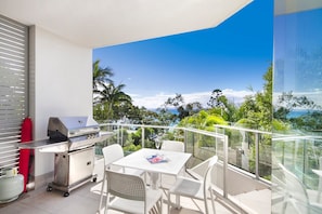 Un balcon équipé d’un barbecue, d’une table à manger, de chaises et offrant une vue sur l’océan et la végétation.