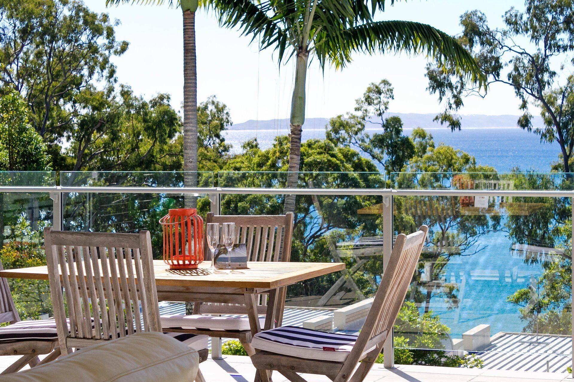 A wooden table with chairs and a glass railing overlooking a pool and ocean view.