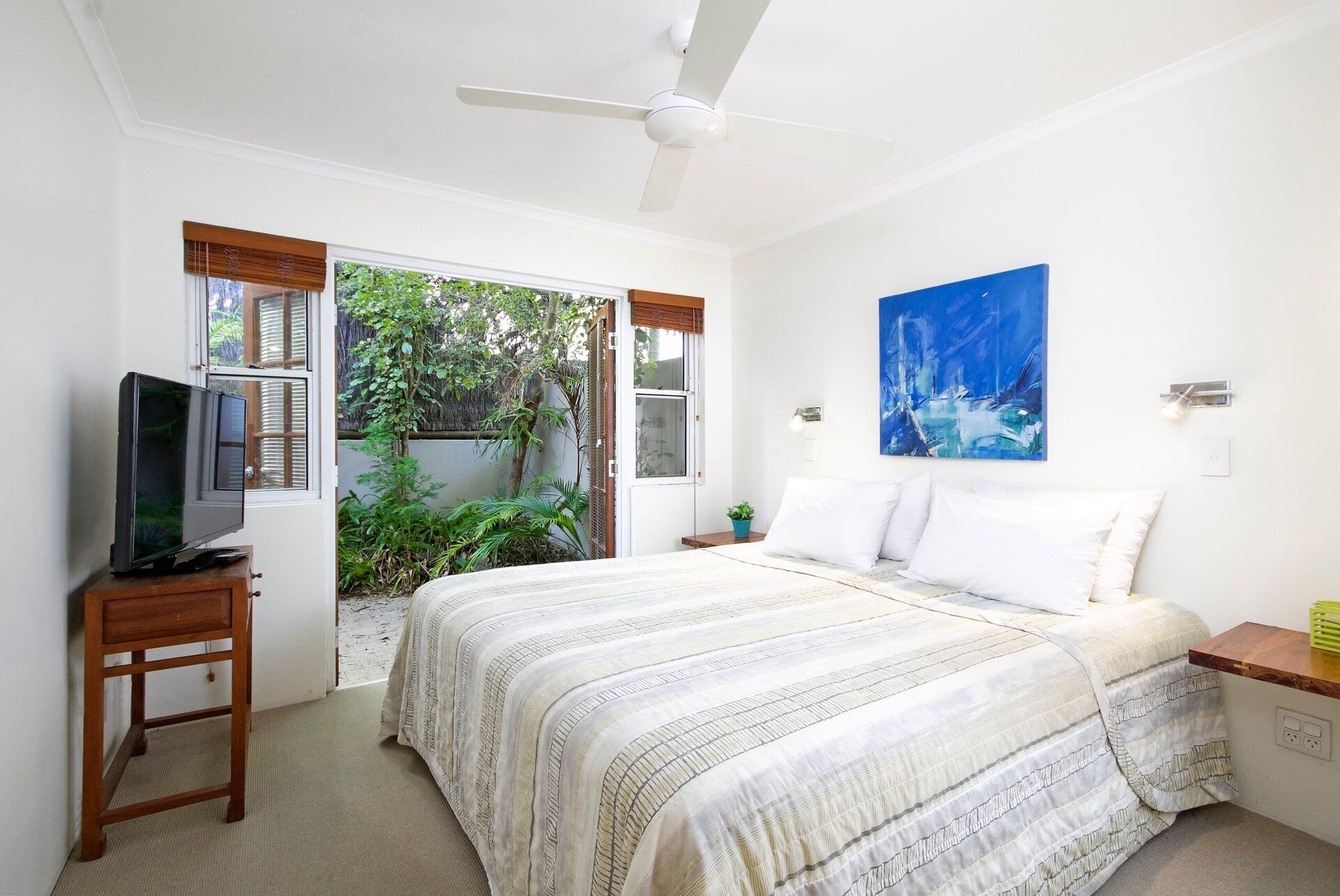 A bedroom with a bed, a television on a wooden stand, a ceiling fan, a window with blinds, a painting, and a small table with a plant.