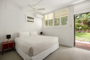 A bedroom with a bed, a nightstand, a ceiling fan, a window with blinds, and a door leading to a garden. - Bayona 2 - Family Beach Charm (Little Cove)