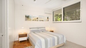 A bedroom with a bed, two bedside tables, a window with blinds, and wall-mounted artwork. - Bayona 4 - Balcony Tranquility (Little Cove)