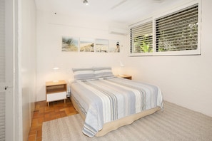 Une chambre à coucher comprenant un lit, deux tables de chevet, une fenêtre avec des stores et des œuvres d’art accrochées au mur.