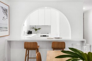 Cozinha moderna com bancada branca, duas cadeiras de madeira, uma mesa de jantar e um espelho grande em arco.