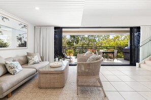 A modern living room with a sofa, coffee table, and wicker chairs, leading to an outdoor patio with a dining set and a grill.