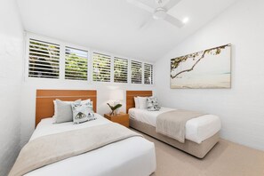 Two single beds with white linens, a ceiling fan, and a framed beach scene adorn this serene hotel room.