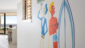 A hallway with a mural of two people on surfboards, a sign reading 'The Junction Hastings St,' and a window with blinds. - Penthouse with Incredible Views & Pool (Noosa Heads)