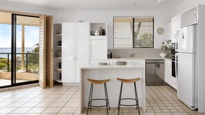 A modern kitchen with white cabinetry, a built-in oven, a stainless steel dishwasher, and a large window with blinds. - Penthouse with Incredible Views & Pool (Noosa Heads)