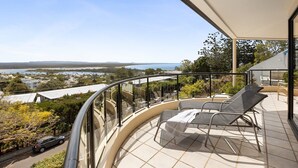 A balcony with a curved glass railing overlooking a coastal landscape with a view of the sea and distant hills. - Penthouse with Incredible Views & Pool (Noosa Heads)