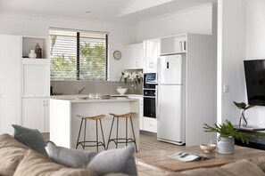 Een moderne keuken met witte kasten, een ingebouwde oven en een koelkast. Er is een raam met zonwering, een spoelbak en een vaas met bloemen op het aanrecht.