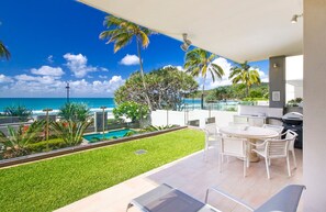 A balcony with a view of the ocean, a pool, and a dining area with outdoor furniture.
