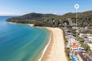 Una vista aerea di una zona costiera con una spiaggia sabbiosa, un mare blu cristallino e una collina verdeggiante sullo sfondo.