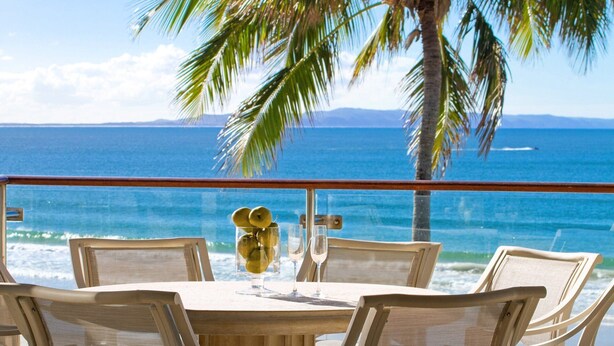 Une table entourée de chaises sur un balcon donnant sur une plage bordée de palmiers et sur l’océan.