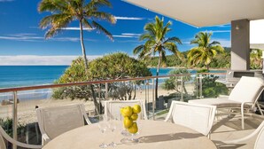 Un balcon doté d’une balustrade en verre, offrant une vue sur une plage et l’océan, avec une table, des chaises et un vase rempli de citrons.
