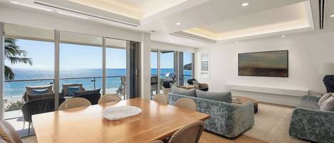 Ein modernes Wohnzimmer mit einem Holzesstisch, Korbstühlen und einer Couch mit Kissen. Ein großes Fenster bietet Blick auf das Meer.