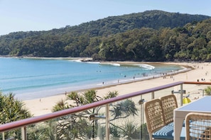 A beach with clear blue water, a sandy shore, and a forested hill in the background.