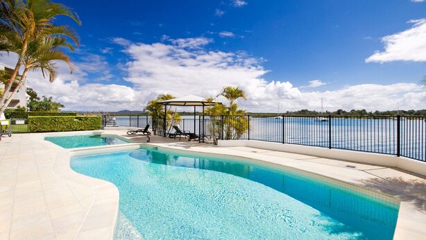 Une piscine au design incurvé, entourée d’une terrasse carrelée de couleur blanche, d’un espace de détente abrité avec des chaises longues, et offrant une vue sur une étendue d’eau dont on aperçoit la rive au loin.