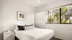 Un dormitorio con cama, mesita de noche, lámpara de noche, ventana con vista a árboles y un cuadro en la pared.