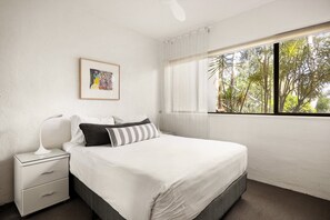 A bedroom with a bed, a nightstand, a bedside lamp, a window with a view of trees, and a framed picture on the wall.