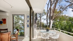 Un balcón con mesa y sillas, vistas a árboles y una masa de agua, y una puerta que da a un espacio interior con una silla y una cesta.