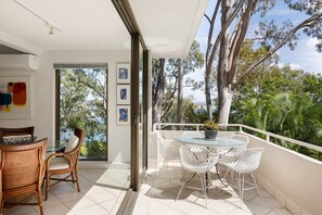 A balcony with a dining table and chairs, a view of trees and a body of water, and a door leading to an interior space with a chair and a basket.