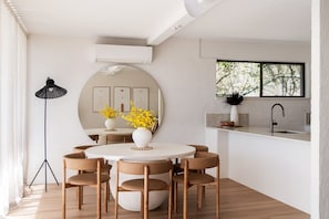A modern dining area with a round table, six chairs, a mirror, and a window with a view of trees.