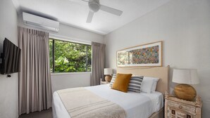 A hotel room with a bed, a ceiling fan, a window with curtains, a wall-mounted TV, a bedside table with a lamp, and a decorative wall piece. - Cove Point 5 - Stylish Cove Escape (Noosa Heads)