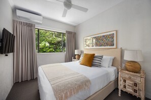 A hotel room with a bed, a ceiling fan, a window with curtains, a wall-mounted TV, a bedside table with a lamp, and a decorative wall piece. - Cove Point 5 - Stylish Cove Escape (Noosa Heads)
