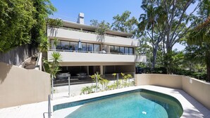 A two-story building with a swimming pool, surrounded by trees and a clear sky. - Cove Point 5 - Stylish Cove Escape (Noosa Heads)