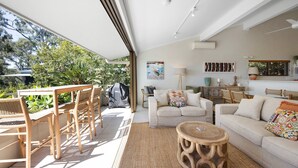 A spacious living area with a sofa, chairs, and a coffee table. There is a grill outside and a view of greenery. - Cove Point 5 - Stylish Cove Escape (Noosa Heads)
