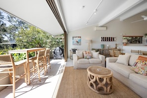 Een ruime woonkamer met een bank, stoelen en een salontafel. Buiten is er een barbecue en uitzicht op groen.