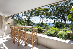 A balcony with a table and chairs, overlooking a coastal landscape with trees and the sea. - Cove Point 5 - Stylish Cove Escape (Noosa Heads)