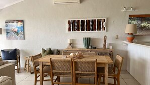 A dining area with a wooden table and wicker chairs, a painting on the wall, and a lamp on the counter. - Cove Point 5 - Stylish Cove Escape (Noosa Heads)