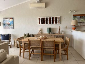 Een eetgedeelte met een houten tafel, rieten stoelen, een schilderij aan de muur en een lamp op het aanrecht.