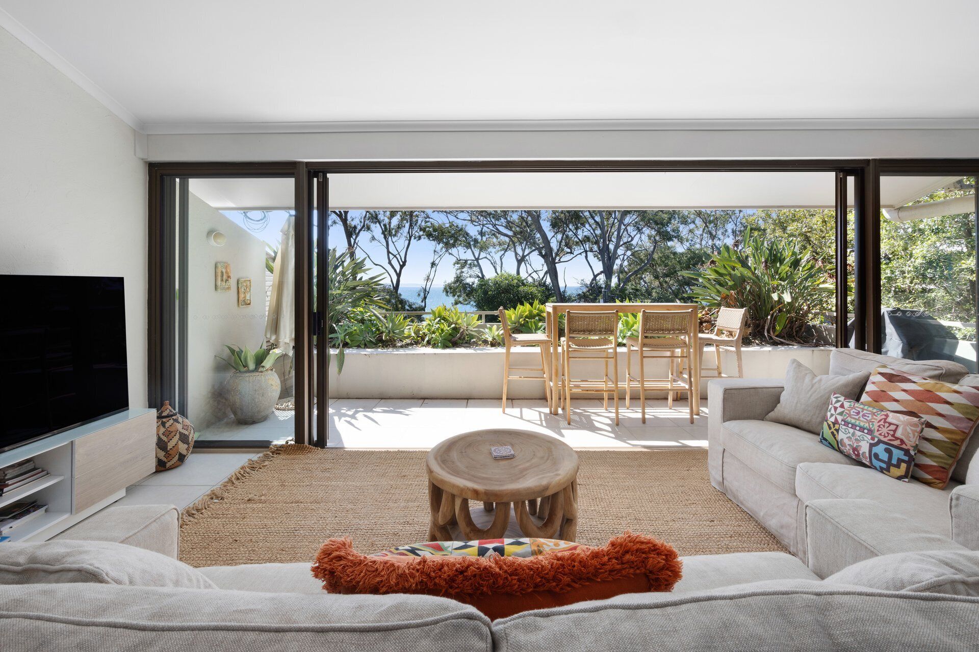 A modern living room with a flat-screen TV, a sofa, a coffee table, and a view of the outdoors through large glass doors.