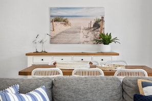 Un comedor con una mesa de madera, sillas de mimbre blanco, un sofá blanco con cojines a rayas y un cuadro con temática de playa en la pared.