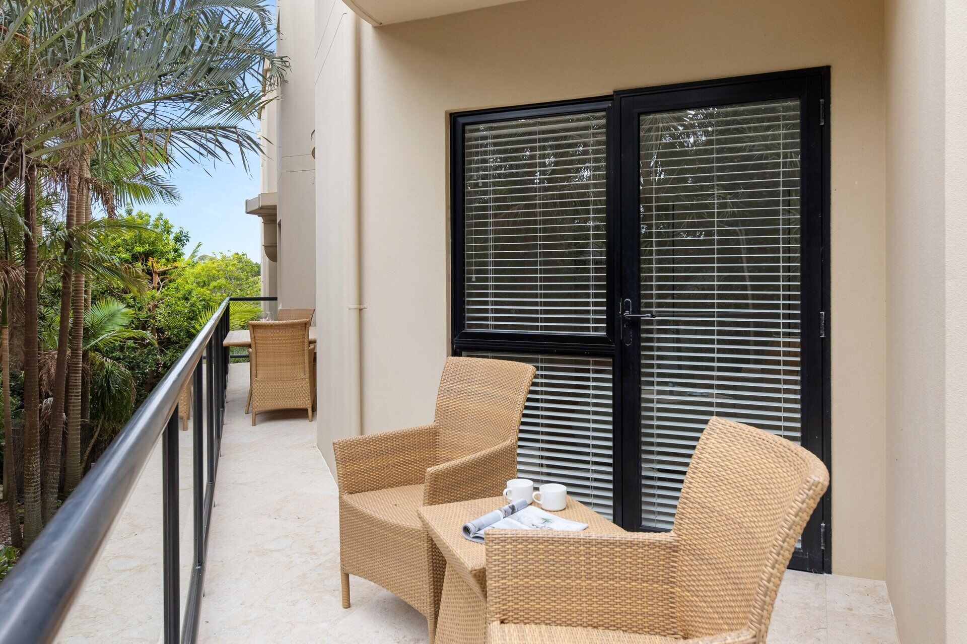A balcony with wicker furniture, a door with blinds, and a view of greenery.